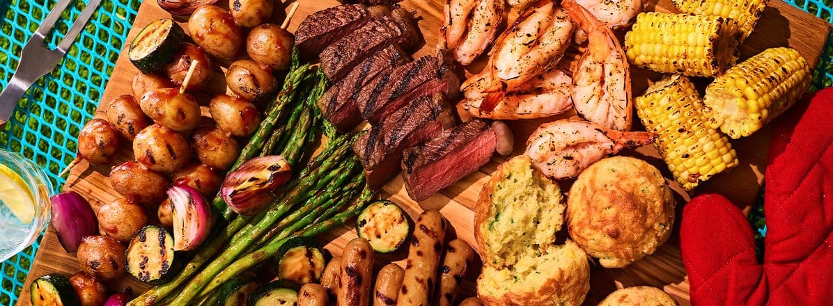 Overhead shot of BBQ-uterie board with sliced tri tip streak, grilled asparagus, corn bread, sausage, potato, shrimp, and corn. 