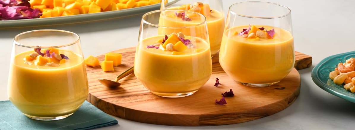 Four glasses of Mango Pudding with chunks of mango, cashews and dried roses as garnish and also on plates around the glasses.