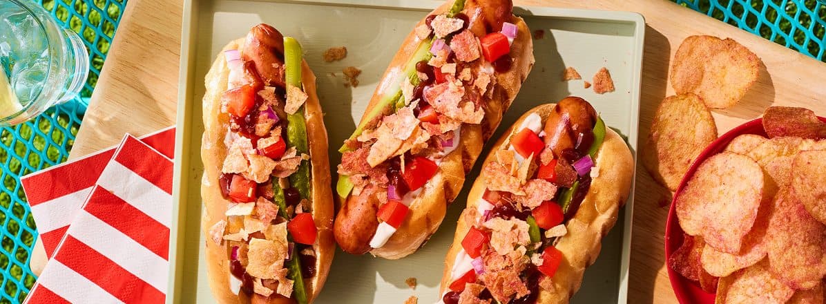 Grilled canadian hot dog with ketchup, crushed ketchup chips, bbq sauce, sour cream, tomato, red onion and crushed ketchup chips