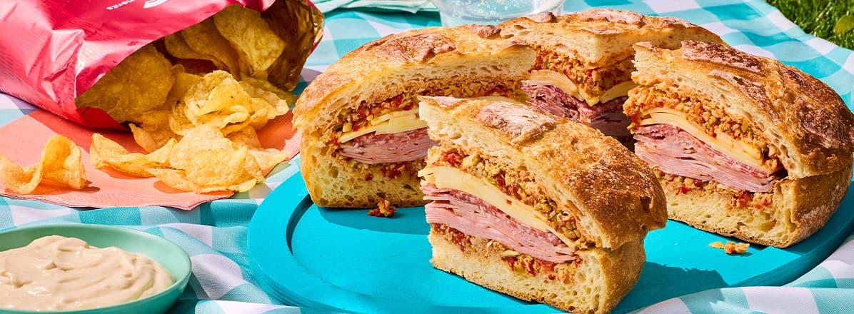 large blanket with a sliced muffuletta sandwich bag of chips and side dip with sparkling clear drinks in the background