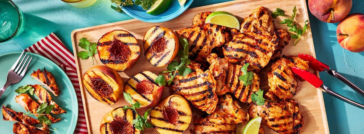 Bamboo cutting board with red tongs, grilled peaches and curried chicken thighs ready to serve