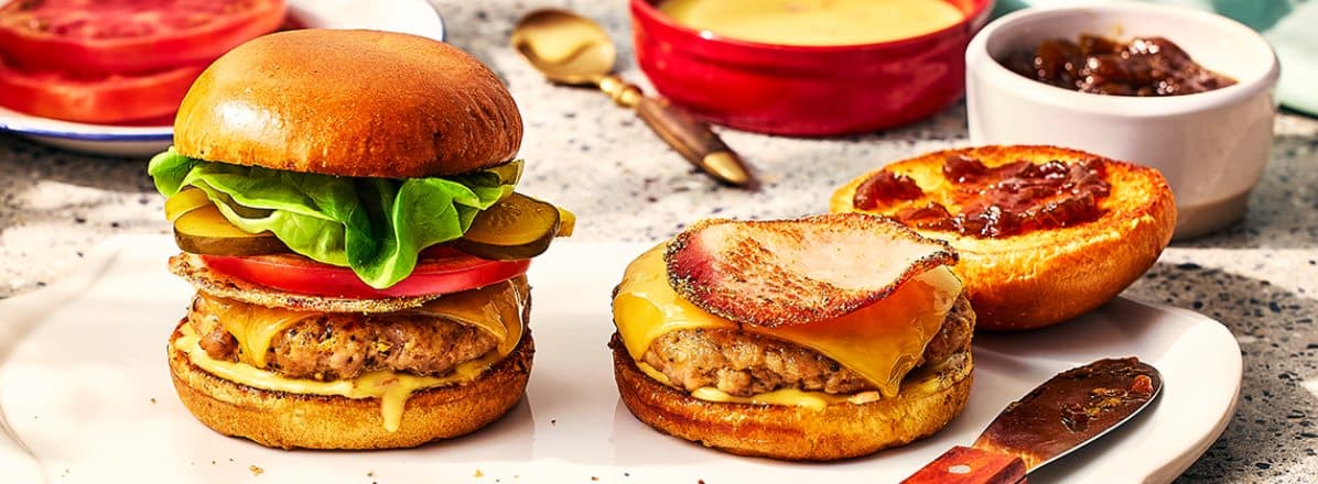 Pork and peameal bacon burger stacked high in a brioche bun, topped with pickles, tomato and cheddar cheese.