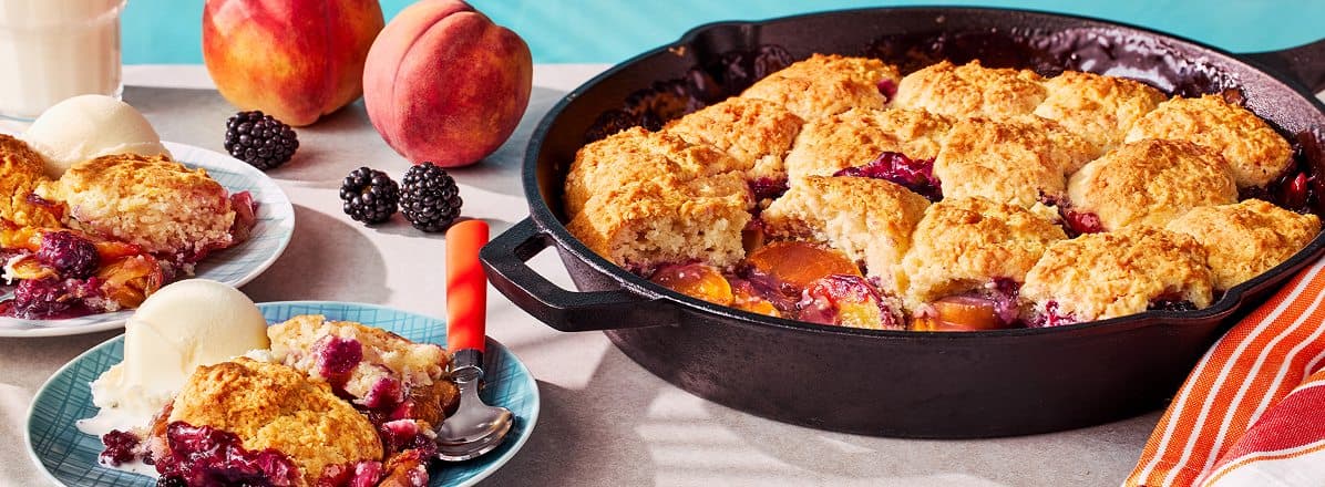 large cast iron pan of Grilled Peach and Blackberry Cobbler, with two servings a la mode to the left on side plates