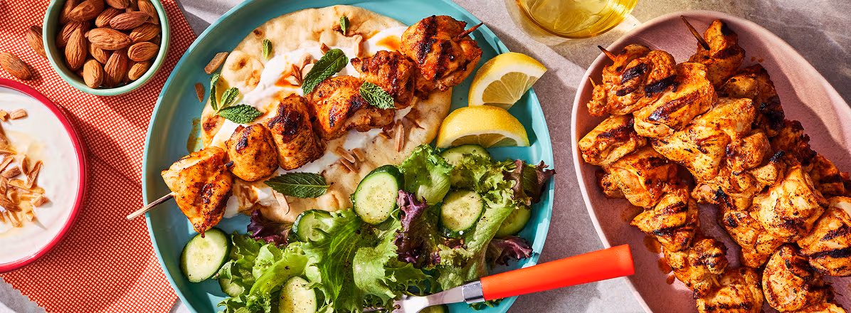 pink dinner plate with harissa grilled chicken skewer over yogurt sauce and naan bread with side green salad, and side platter of remaining chicken and harissa skewers
