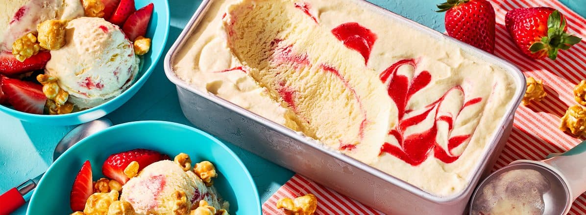 loaf pan of frozen, partially scooped Corn and Strawberry Semifreddo, with two bowls to the left, topped with sliced strawberries and caramel popcorn