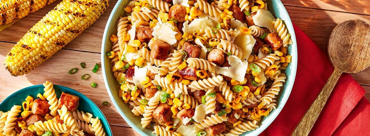 Overhead shot of fusilli pasta salad in a turquoise serving bowl mixed with smoky sausage, charred corn and green onion. Fresh grilled corn to the side.
