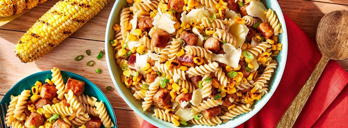 Overhead shot of fusilli pasta salad in a turquoise serving bowl mixed with smoky sausage, charred corn and green onion. Fresh grilled corn to the side.