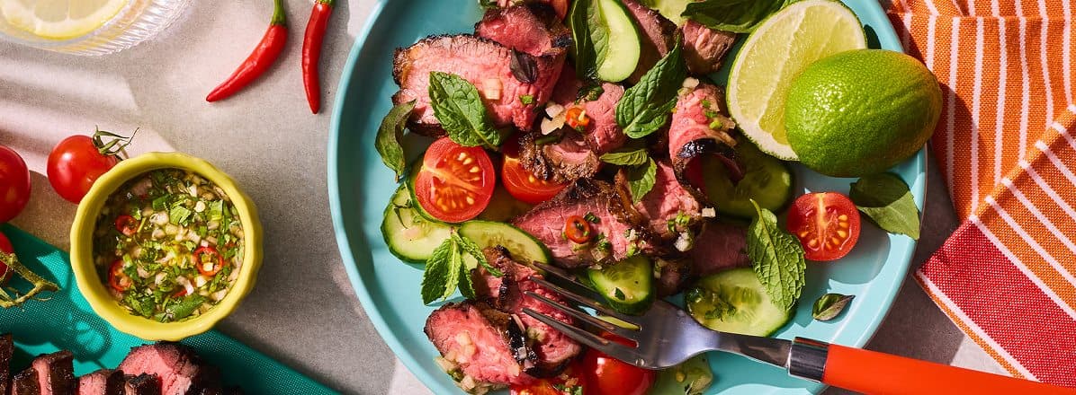 round blue platter of marinated, grilled and sliced crying tiger beef, fresh herbs, lime half, with Nam Jin Jaew sauce on the side