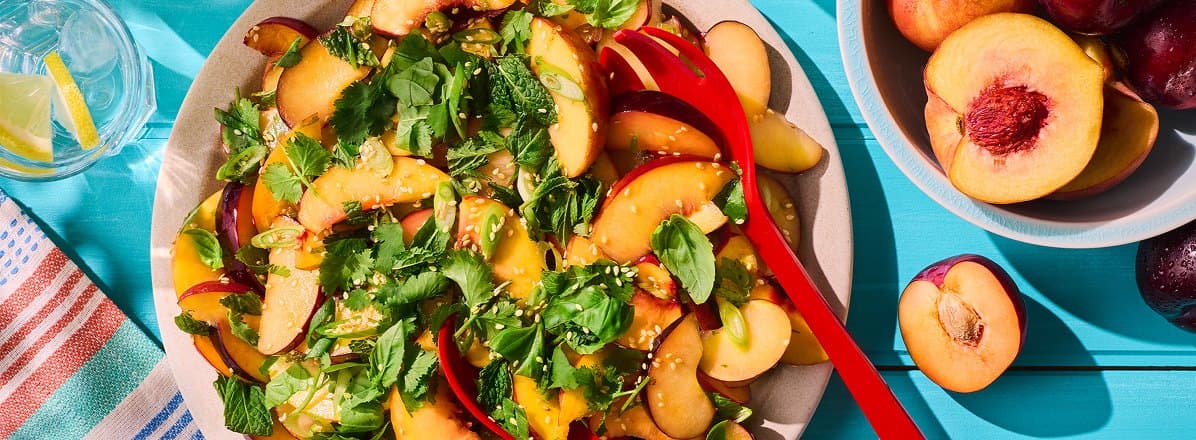 Overhead shot of peach salad topped with herbs, and sesame seeds. Bowl of fresh stone fruit like peaches and plums to the side.