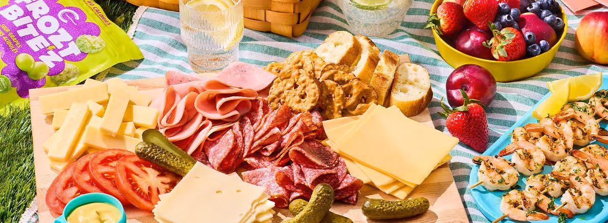 picnic blanket with cutting board topped with Summer Deli cheese & meat board, bowl of fruit, bag of Compliments Frozt Bite’z Frozen Sour Grapes, and Panache Shrimp Skewers - Herb & Garlic skewers.