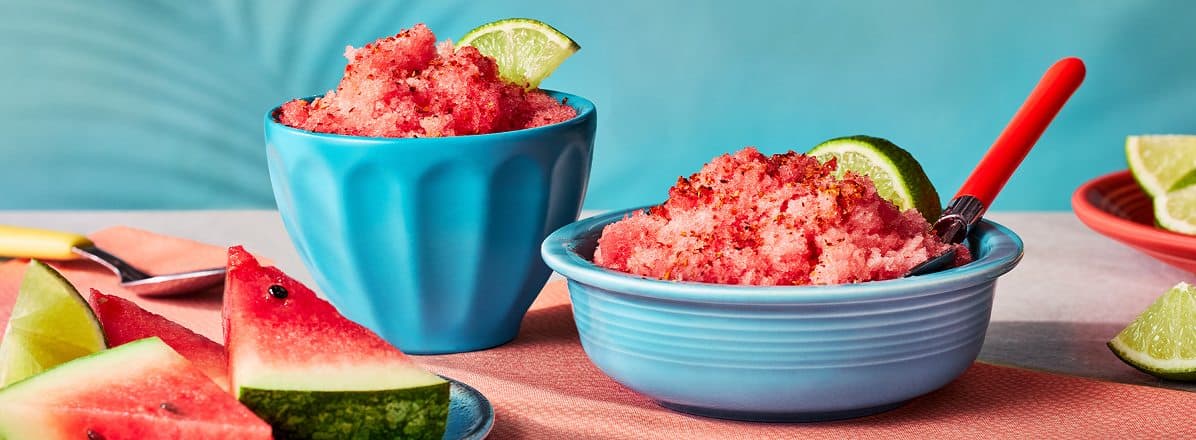 two blue bowls of watermelon shaved ice with tajin and lime garnish, and plate of watermelon triangles to the side