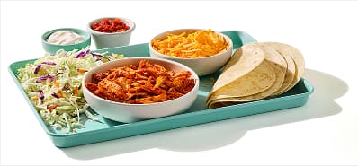 Tray holding a bowl of al pastor chicken, a bowl of shredded cheese, folded tortillas, and a bed of shredded cabbage. On the side, two small dipping bowls of sour cream and salsa.