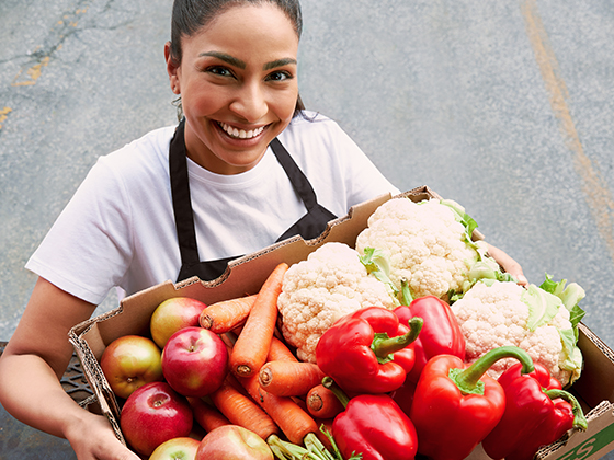 A woman working with Second Harvest is looking up to the camera with a smile on her face as she holds a large cardboard box filled with red peppers, cauliflour, carrots, and apples that have been rescued from waste.