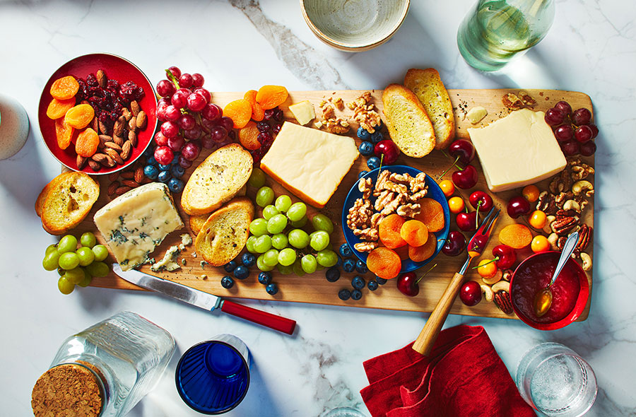 Wooden board with wedges of cheese, nuts, bread, grapes, and dried fruit.