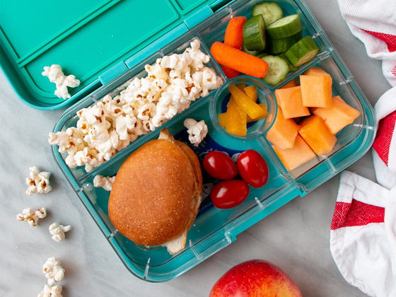 Overhead shot of a packed lunch with sandwich and portion of popcorn, cut fruit and veggies.