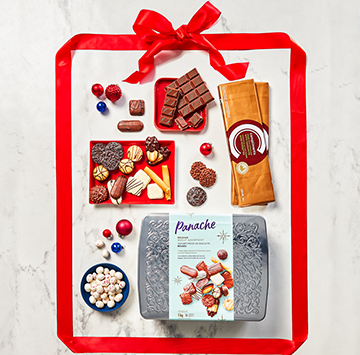 Flat lay on white marble with Panache Luxury Belgian Biscuit Collection, Compliments Peppermint & Dark Chocolate Almonds, and Compliments Swiss Milk Chocolate with Hazelnuts, and a red ribbon wrapped around it all