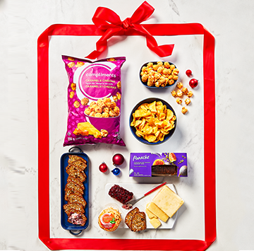 Flat lay on marble with Panache Honey Dijon Flavour Kettle Cooked Potato Chips, Compliments Caramel & Cheddar Popcorn and Cheeseboard gift pack with Red ribbon Around it all