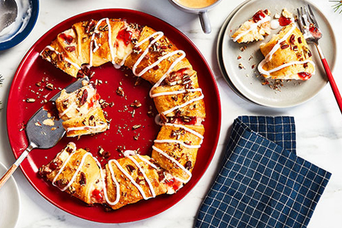 Strawberry crescent roll wreath with an icing sugar glaze drizzle, on a red serving platter with mugs of coffee and side plates.