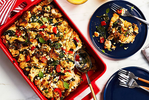 Mushroom, spinach and goat cheese strata in red casserole dish with dark blue serving plates on the side.