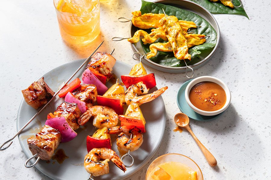 Three Pan-Asian style skewers including chicken, pork belly and shrimp on green-lined platter and a gray platter with orange beverage to one side.