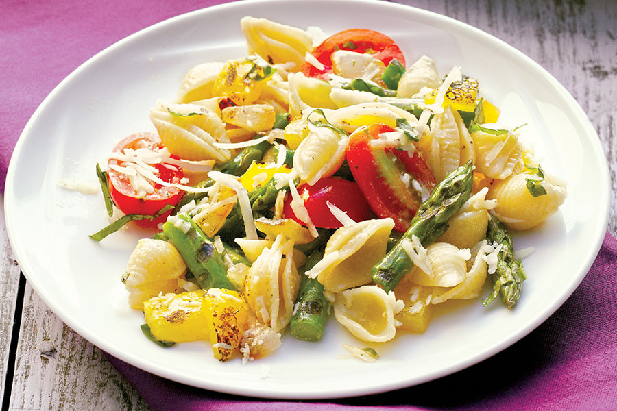 White plate of asparagus pasta with fresh parmesan and grape tomatoes on table with purple napkin.