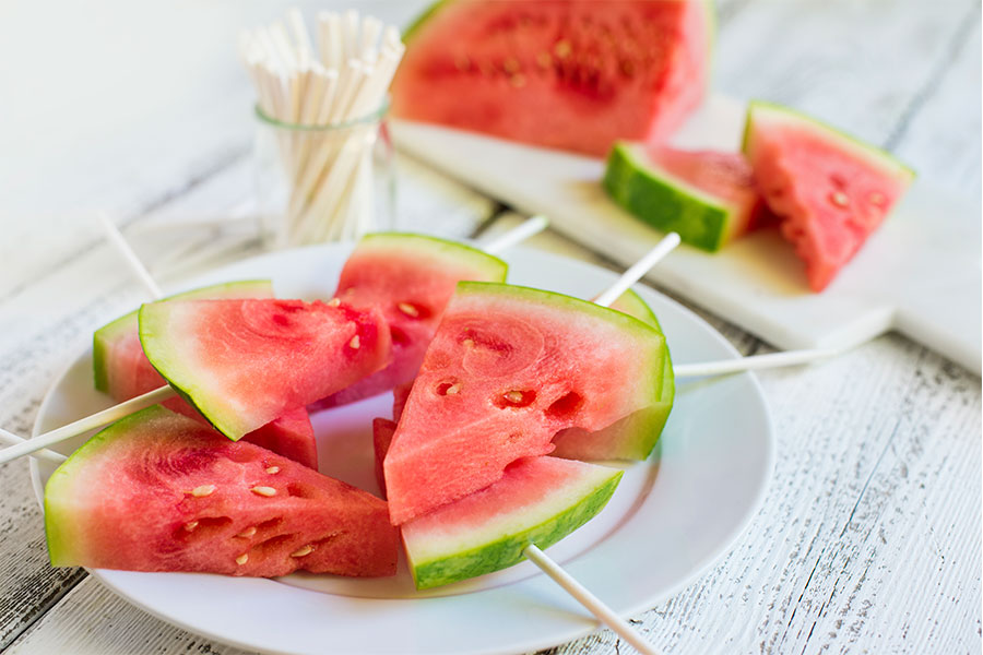 A white plate of watermelon slices cut out as triangles with ice-pop sticks stuck in the end.