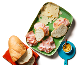 Italian cold cuts, baguette bread slices, and a wheel of Brie cheese on a blue serving platter next to a little green plate with bread on it.