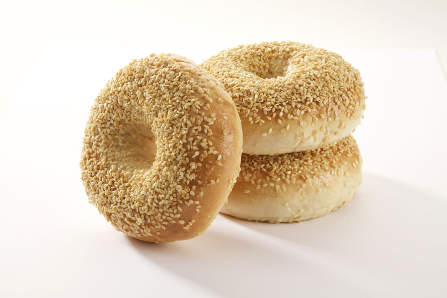 Three sesame seed bagels, two stacked on top of each other with one leaning against it on a white board.