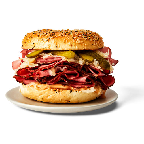 Everything Reuben bagel including sliced pastrami, sauerkraut, Swiss cheese slices, and pickles on a white plate.