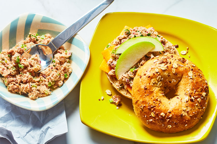 Multigrain bagel topped with a tuna salad, melted cheese and apple slices on a bright yellow plate.