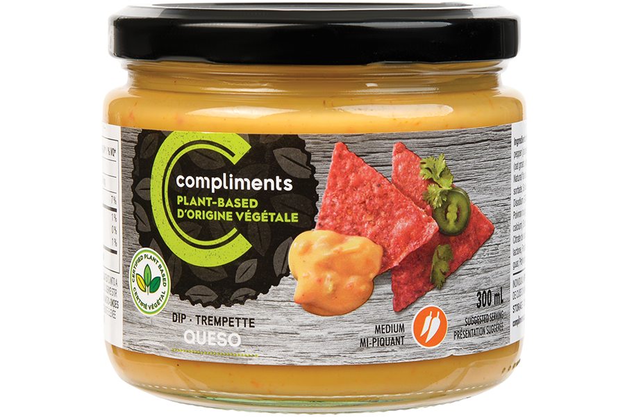 A clear glass jar of Compliments Plant-Based Queso Dip pictured as a dip alongside red tortilla chips on the jar artwork.