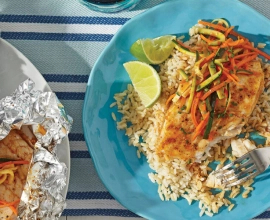 Pickerel fish fillets and vegetables wrapped in tinfoil beside a piece of fish topped with vegetables on a bed of rice sitting on a blue plate.