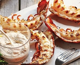 Grilled lobster tails on a platter beside a cup of aioli with porcelain spoon beside two lemon wedges