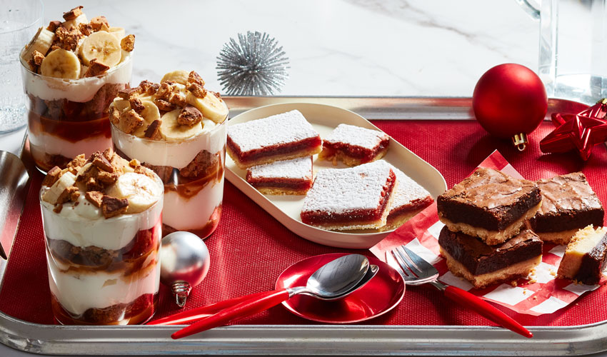White marble counter with holiday decor in the background, and a tray of three holiday desserts front and center: Banana Bread Banoffee trifle, Cranberry Squares and Brownie Shortbread Bars.
