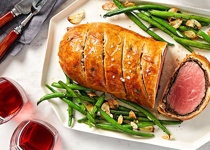 Top-down view of table with serving dish of beef wellington with green beans and almonds on either side. Main dish beside two glasses of red liquid, plate of slice of wellington and beans, plate with ramekin of mustard and ramekin of salt.