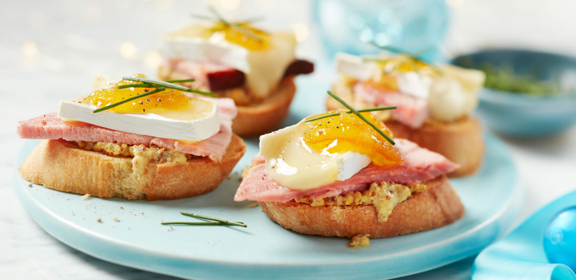 Light blue round plate with four ham and brie-topped crostinis topped with marmalade and fresh chives.