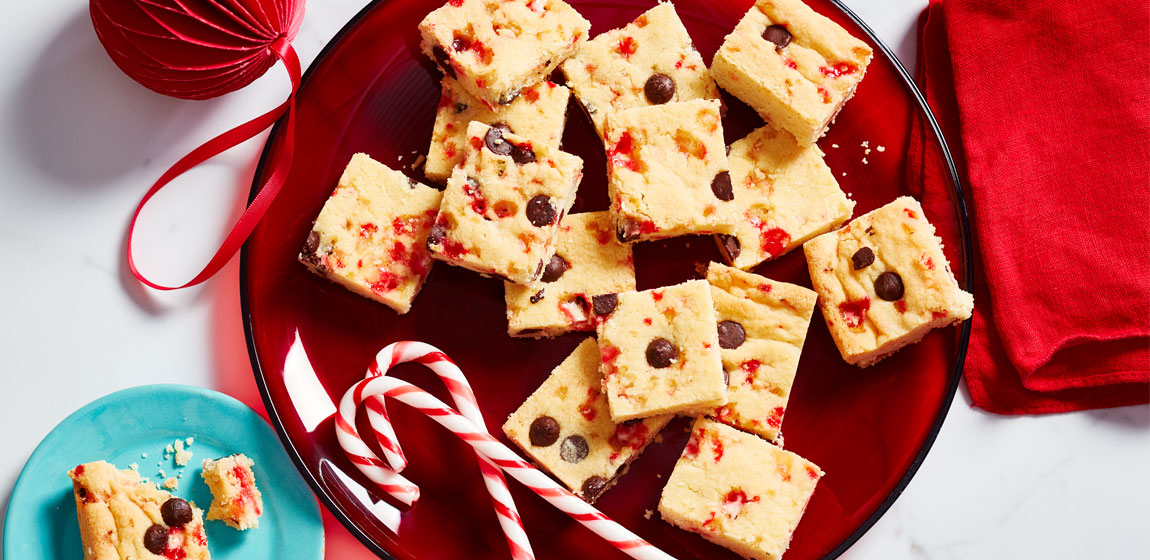 White marble tabletop with holiday decorations and a red plate of Cherry Chip shortbread squares and two candy canes.