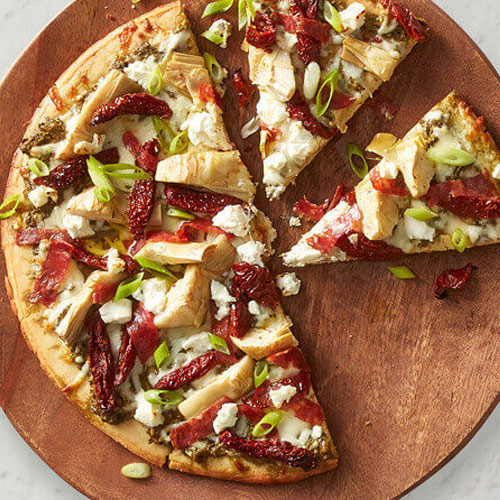 Grilled naan bread topped pizzas with pesto, mozzarella, sundried tomatoes, calabrese sausage, artichoke hearts, green onion and goat cheese on a white speckled plate and marble and wooden cutting board.