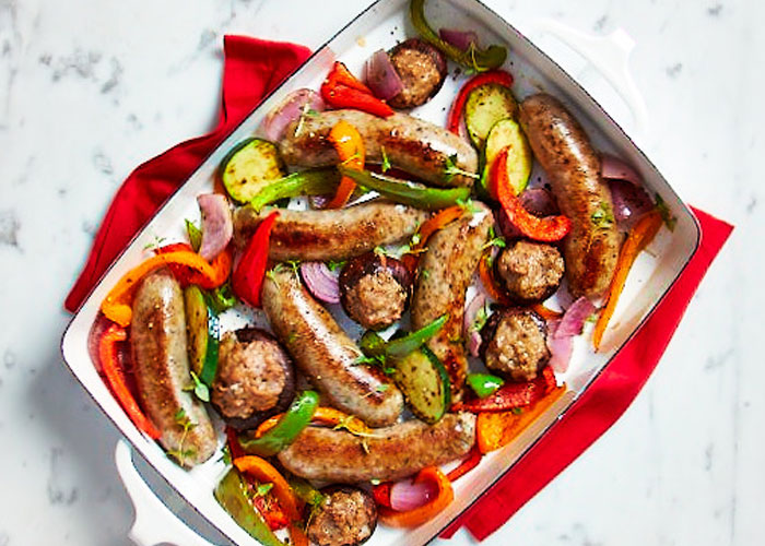 White casserole dish with festive holiday sausage with mixed roasted vegetables on a red tea towl over a white countertop.