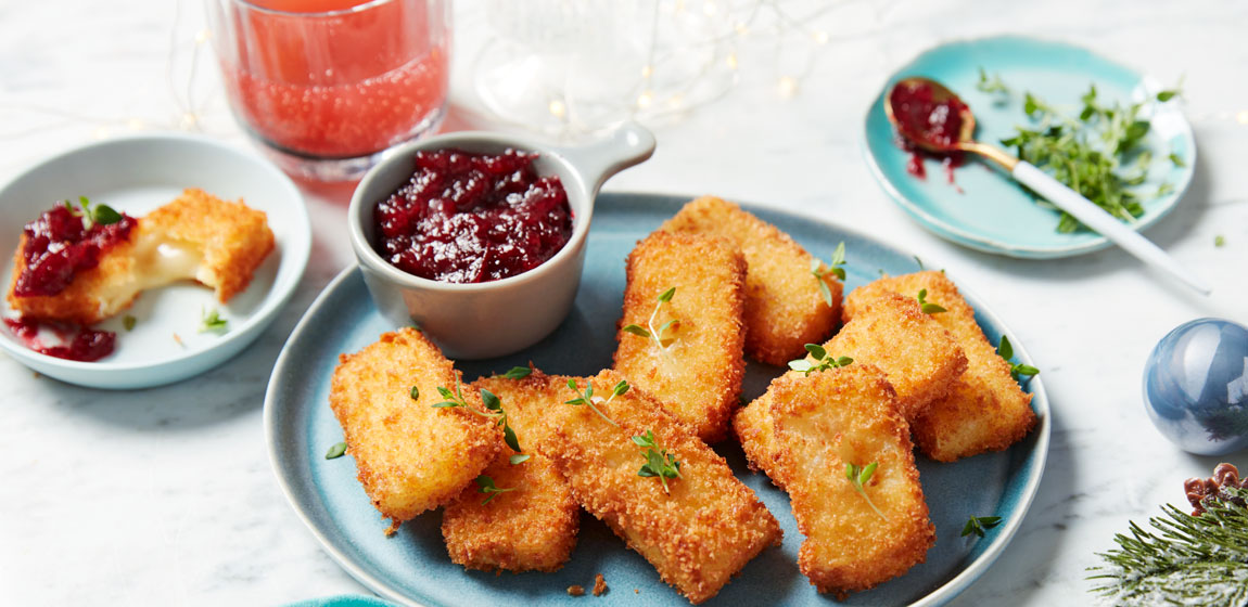 Light blue round plate of fried brie slices and a serving dish of Panache Cranberry and Port Topper and Spread on top, set on a white marble tabletop next to a napkin, with holiday decorations in the background.