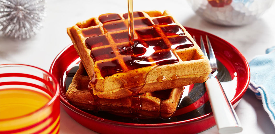 Close-up of waffles on a red plate with syrup being drizzled down from up high, and a fork on the side.