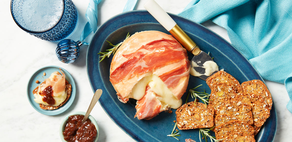 Large oval blue platter with a serving knife, crackers and a wheel of prosciutto-wrapped brie, set on a while marble tabletop with holiday decorations.