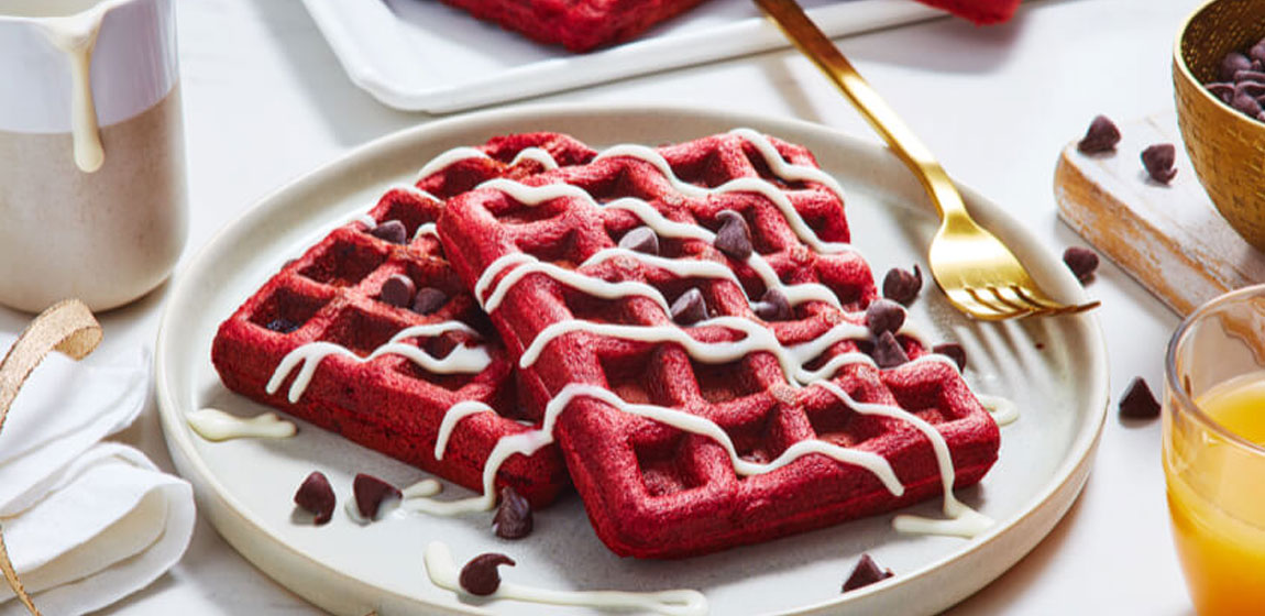 Red velvet waffles topped with white icing and chocolate chips on a white plate with a gold fork.