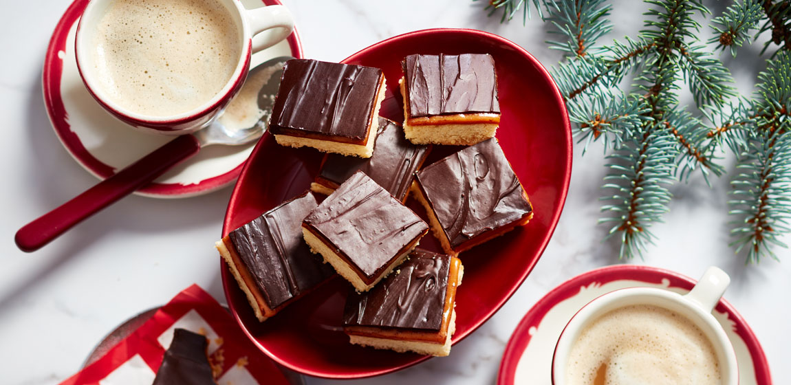 white marble tabletop with oval red plate of millionaire shortbread bars and holiday decorations on the table around it.