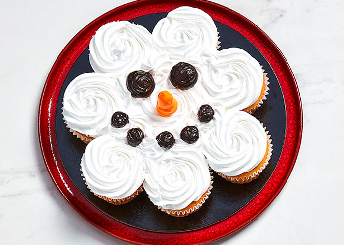 White marble surface with a red plate of cupcakes with a single snowman face designed into the icing of them all.