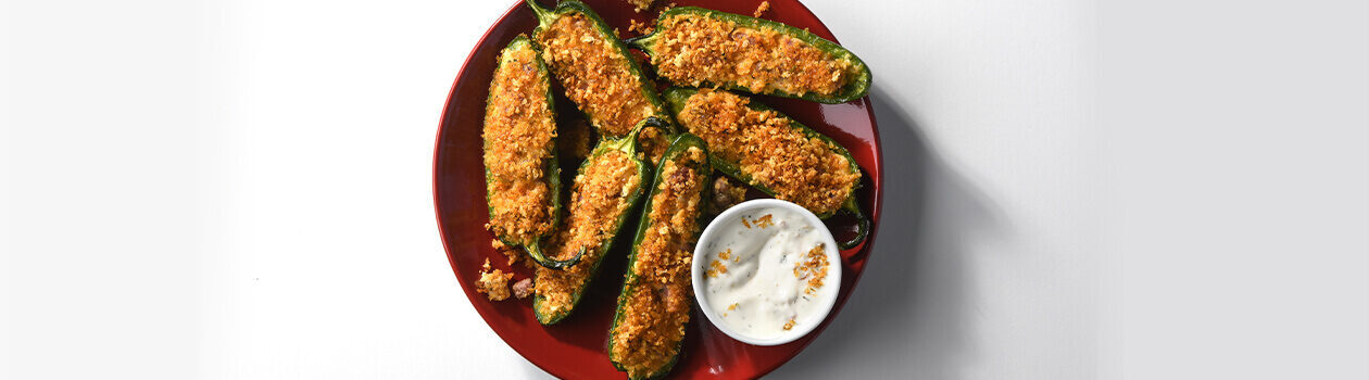 red plate with several crispy meat & cheese jalapeno poppers on top with a ramekin of dipping sauce.