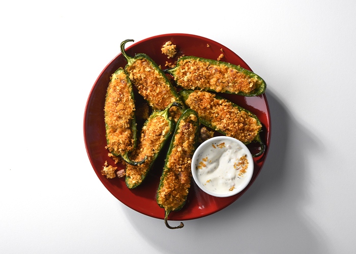 Red plate with several crispy meat & cheese jalapeno poppers on top with a ramekin of dipping sauce.