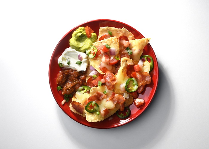 Red side plate full of nachos, melted cheese, sausage pieces, jalapeno pepper rounds and red onion, sour cream, guacamole and salsa