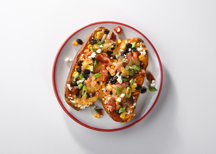 White, red rimmed plate with loaded baked sweet potatoes scattered with cheese, black olive pieces, green onions and more.