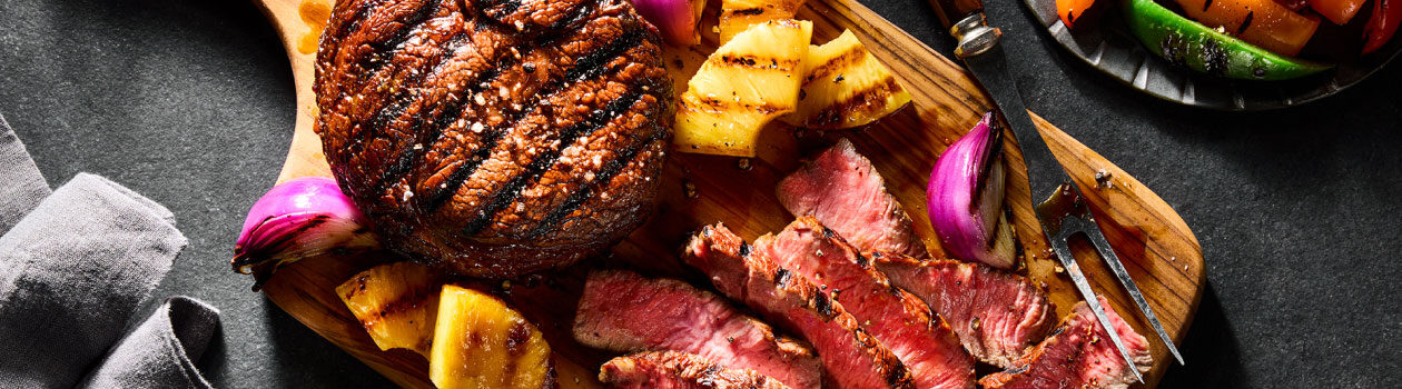 Grilled Hawaiian Ribeye Steak on a wooden cutting board next to slices of grilled pineapple and red onion quarters.